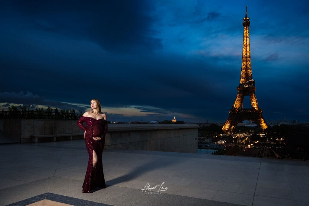 Paris Babymoon photography image of pregnant woman in a red dress stood beside the Eiffel Tower in the night.